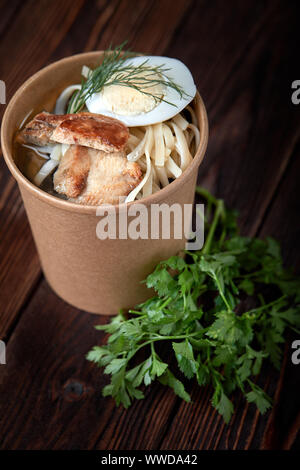 Pappbecher mit Nudeln mit chiken und Eier auf Holzuntergrund mit Kräutern Stockfoto