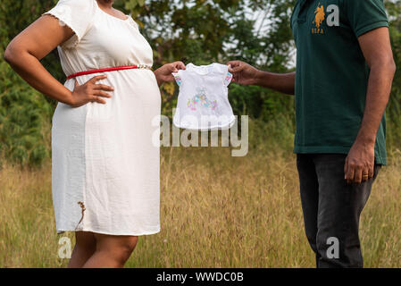 Schwangere Frau und ihr Ehemann Holding Baby Kleidung und zeigt ihren Babybauch an einer im Schießen Stockfoto