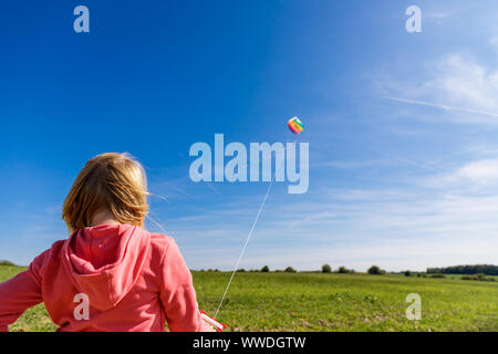 Hübsches kleines Mädchen mit blondem Haar in einem Feld in einem rosa sweatshirt blickt auf eine bunte Drachen in einem blauen Himmel mit weichen Wolken Stockfoto