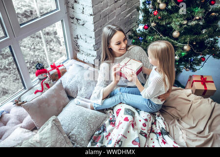 Fröhliche Mamma und ihre süße Tochter Mädchen den Austausch von Geschenken in der Nähe der Fenster. Eltern und Kind Spaß haben in der Nähe von Weihnachten Baum drinnen. Morgen Weihnachten. Stockfoto