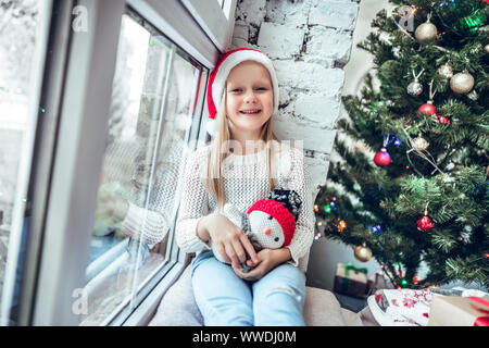 Portrait von niedlichen glückliche kleine Mädchen mit Weihnachten Red hat. Lustig Kind sitzt auf der Fensterbank, festlich mit Holiday details dekoriert Stockfoto