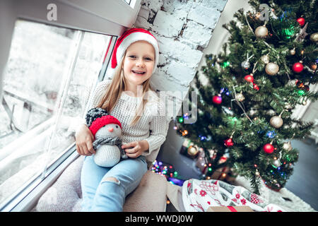 Süßes kleines Mädchen sitzt am Fenster und schaut in die Kamera. Zimmer dekoriert auf Weihnachten. Stockfoto