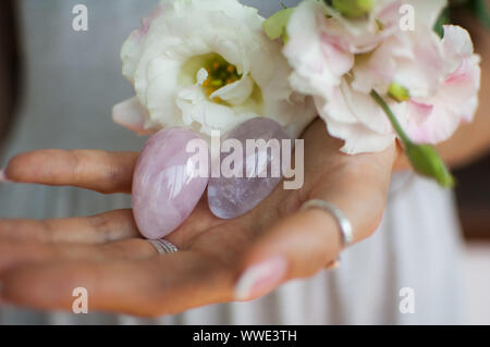 Weibliche Hände halten zwei Yoni Eier für vumfit, imbuilding oder Meditation werden aus Rosenquarz und Amethyst violett transparent mit weißen Blumen gemacht Stockfoto