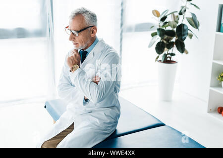 Nachdenklich bärtigen Doktor in Gläsern und weißes Fell sitzen auf der Massageliege und berührt, Gesicht Stockfoto