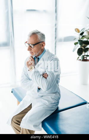 Nachdenklich bärtigen Doktor in Gläsern und weißes Fell sitzen auf der Massageliege und berührt, Gesicht Stockfoto