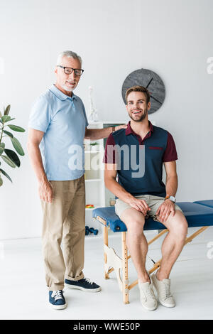 Positive bärtigen Mann in Gläsern in der Nähe von Happy stehenden Patienten in der Klinik Stockfoto