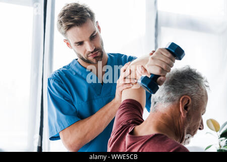 Selektiver Fokus der Arzt in der Nähe der mittleren gealterten Mann Trainieren mit hantel Stockfoto