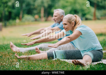 Lächelnd reifer Mann und Frau praktizieren Stretching stellt beim Sitzen auf Yoga Matten in Park Stockfoto