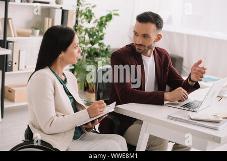 Behinderte Unternehmerin schreiben in Zwischenablage während am Schreibtisch sitzt und spricht mit Business Partner Stockfoto