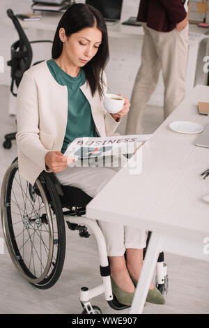 Attraktive deaktiviert Geschäftsfrau lesen Zeitung und halten Kaffee Tasse beim Sitzen im Rollstuhl am Arbeitsplatz Stockfoto