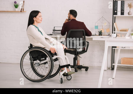 Lächelnd deaktiviert Geschäftsfrau weg schauen beim Sitzen im Rollstuhl in der Nähe von Kollegen Stockfoto