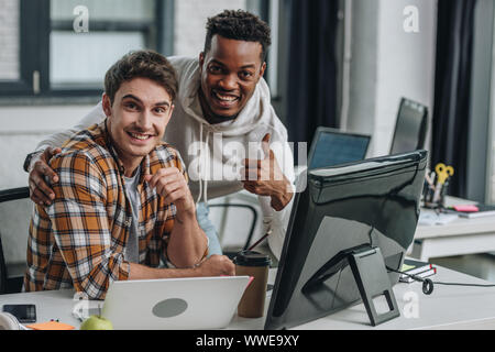 Zwei fröhliche multikulturelle Programmierer auf Kamera und zeigen Daumen hoch Stockfoto