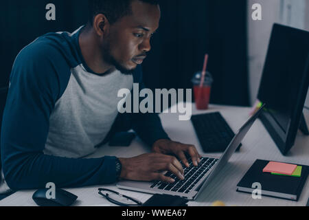 Aufmerksame afrikanische amerikanische Programmierer mit Laptop, während in der Nacht in die Arbeit im Büro Stockfoto