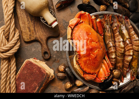 Blick von oben auf die Ungekocht, Krabben, Muscheln, Herzmuscheln und Miesmuscheln in der Schüssel in der Nähe von Krümelig altes Buch, Seil und Flasche mit Korken auf Holz Schneidebrett auf Textur Stockfoto