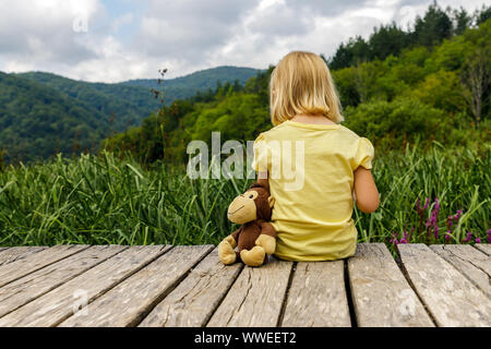 Kleines Mädchen mit Affe sitzt auf Holzsteg und Träume. Tourismus. Aktive Familie Zeit in der Natur. Wandern mit Kindern Stockfoto