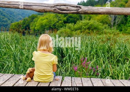 Kleines Mädchen mit Affe sitzt auf Holzsteg und Träume. Tourismus. Aktive Familie Zeit in der Natur. Wandern mit Kindern Stockfoto