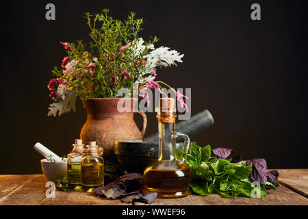 Ton Vase mit frischen Kräutern und Blumen in der Nähe von Mörser und Stößel und Flaschen auf Holztisch isoliert auf Schwarz Stockfoto