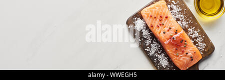 Blick von oben auf die rohen Lachs Steak mit Pfefferkörnern und Salz auf hölzernen Schneidebrett in der Nähe von Öl auf Marmor Oberfläche, Panorama-Aufnahme ein. Stockfoto