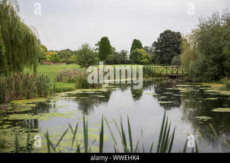 Großer Teich mit Spiegelungen der Bäume und der Himmel im ruhigen Wasser des Teiches, mit einer kleinen Holzbrücke über das Ende der Teich Stockfoto