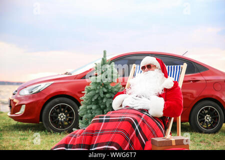 Weihnachtsmann mit Tannenbaum und Geschenke sitzen auf Stuhl in der Nähe von Auto auf Riverside Stockfoto