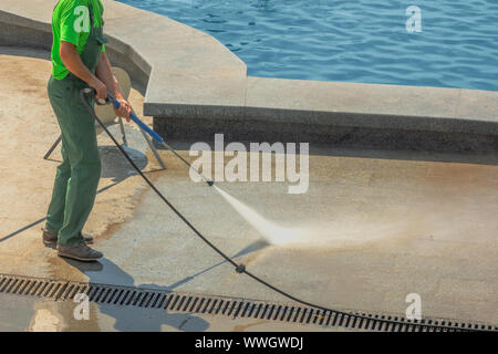 Arbeitnehmer Mann in Uniform wäscht Street oder Park Bürgersteig in der Nähe von Wasser Pool oder Brunnen. Kommunale Dienste der Stadt Reinigung. Kerl Spritzausrüstung verwendet. Stockfoto