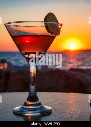 Malerische Ansicht aus Glas mit alkoholischen Getränken und die Scheibe von Kalk auf Tisch gegen bunte Himmel über dem Meer bei Sonnenuntergang Stockfoto