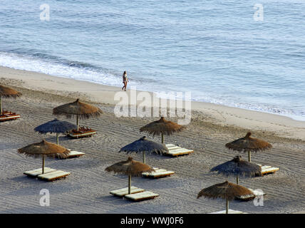 Lonely Beach Walker, der am Ufer des Meeres vor der strohgedeckten Sonnenschirmen und Liegestühlen Stockfoto