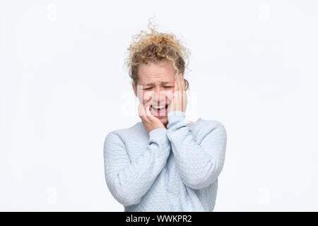 Frustrierte junge blonde lockige Frau in Blau Pullover in schreckliche Kopfschmerzen die Hände auf den Kopf finsteres Gesicht mit Schmerzen. Studio shot Stockfoto