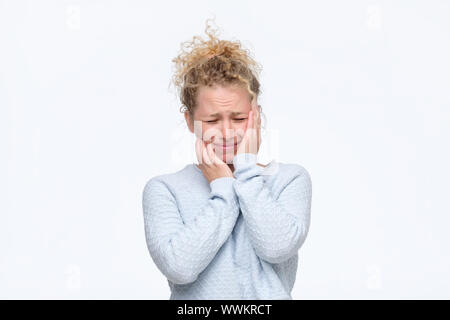 Frustrierte junge blonde lockige Frau in Blau Pullover in schreckliche Kopfschmerzen die Hände auf den Kopf finsteres Gesicht mit Schmerzen. Studio shot Stockfoto