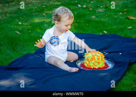 Ein adorable Baby Boy genießt seinen ersten Geburtstag feiern auf einer Party mit einem Kuchen smash eines bunten iced Kuchen draußen im Garten Stockfoto