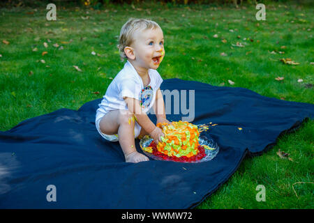 Ein adorable Baby Boy genießt seinen ersten Geburtstag feiern auf einer Party mit einem Kuchen smash eines bunten iced Kuchen draußen im Garten Stockfoto