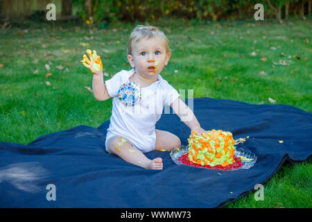 Ein adorable Baby Boy genießt seinen ersten Geburtstag feiern auf einer Party mit einem Kuchen smash eines bunten iced Kuchen draußen im Garten Stockfoto