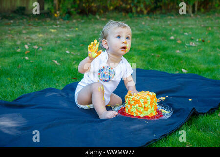 Ein adorable Baby Boy genießt seinen ersten Geburtstag feiern auf einer Party mit einem Kuchen smash eines bunten iced Kuchen draußen im Garten Stockfoto