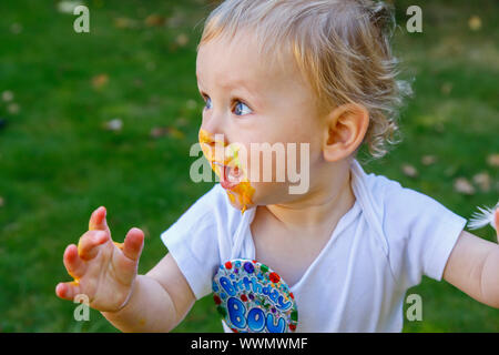 Ein adorable Baby Boy genießt seinen ersten Geburtstag feiern auf einer Party mit einem Kuchen smash eines bunten iced Kuchen draußen im Garten Stockfoto