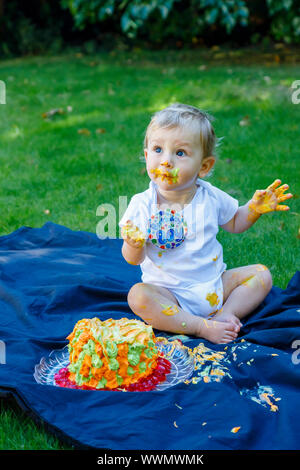 Ein adorable Baby Boy genießt seinen ersten Geburtstag feiern auf einer Party mit einem Kuchen smash eines bunten iced Kuchen draußen im Garten Stockfoto