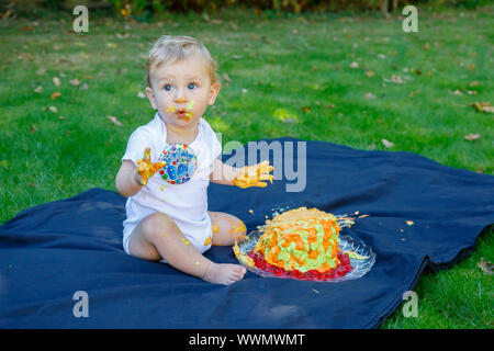 Ein adorable Baby Boy genießt seinen ersten Geburtstag feiern auf einer Party mit einem Kuchen smash eines bunten iced Kuchen draußen im Garten Stockfoto