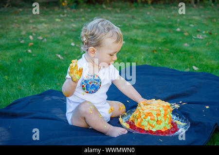 Ein adorable Baby Boy genießt seinen ersten Geburtstag feiern auf einer Party mit einem Kuchen smash eines bunten iced Kuchen draußen im Garten Stockfoto