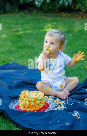 Ein adorable Baby Boy genießt seinen ersten Geburtstag feiern auf einer Party mit einem Kuchen smash eines bunten iced Kuchen draußen im Garten Stockfoto