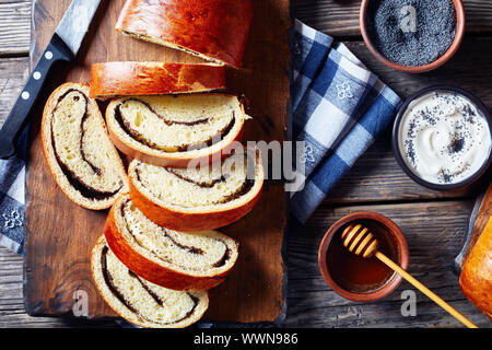 Mohn Kuchen, Makovnjaca, süßem Teig Roulade mit Mohn füllen, in Scheiben geschnitten auf einem Holzbrett, kroatische Küche, Zutaten auf einem rustikalen Tisch, horiz. Stockfoto