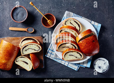 Mohn Kuchen, Makovnjaca, süßem Teig Roulade mit Mohn füllen, in Scheiben geschnitten auf einer weißen Platte auf einer konkreten Tabelle mit Zutaten, kroatische Küche, Stockfoto