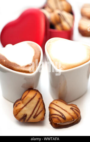 herzförmige Creme Cookies auf rotes Herz Metallbox und paar Cappuccino Kaffeetassen Stockfoto