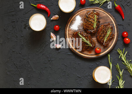 Leckeres barbecue Steaks und Bier in Gläsern auf einem schwarzen Stein, mit Cherry Tomaten und Chili peppers Stockfoto