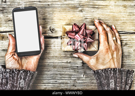 Nahaufnahme von einem Smartphone in den Händen einer Frau und einem Geschenkkarton mit einer roten Schleife auf einen hölzernen Tisch. Stockfoto