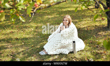 Ein junges Mädchen in einer Merino plaid gewickelt wird durch eine Tasse Tee aus der Thermoskanne auf das Gras in einem Stadtpark erwärmt. Stockfoto