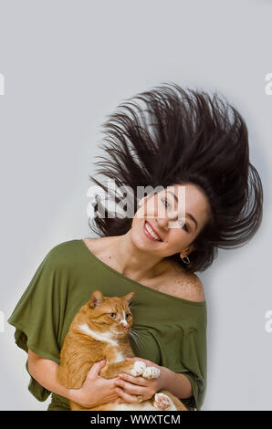 Eine Frau mit grossen Lächeln in Grün shirt ihr dunkles braunes Haar um Schwingen, während sie eine orange Cat, isoliert auf weißem Stockfoto