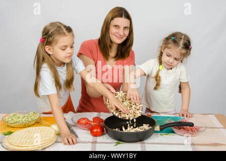 Zwei kleine Mädchen auf dem Küchentisch mit einem Eifer zu helfen, ihre Mutter die Pilze aus der Platte in die Pfanne gießen Stockfoto