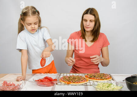 Sechs Jahre altes Mädchen nimmt eine Platte schneiden Tomaten für Pizza unter der Aufsicht von Mama Stockfoto