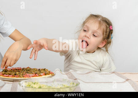 Kleines Mädchen mit Zunge heraus eine Hand um die Pizza zu ziehen, die ihre ältere Schwester bereitet Stockfoto