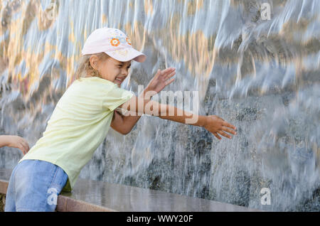 Fröhlich sechs Jahre altes Mädchen im Sommer Kleidung Hand versucht den Wasser künstliche Wasserfall zu erhalten Stockfoto