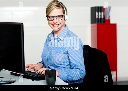 Lächelnde Frau Hilfe der Rezeption Tippen auf Tastatur tragen Kopfhörer mit Mikrofon attached to it. Stockfoto